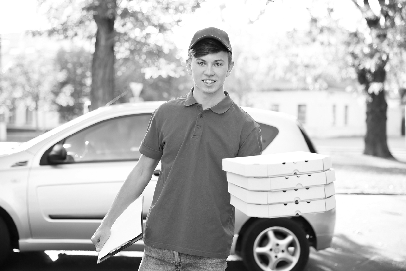 A picture of a pizza delivery boy outside his vehicle holding boxes of pizzas.